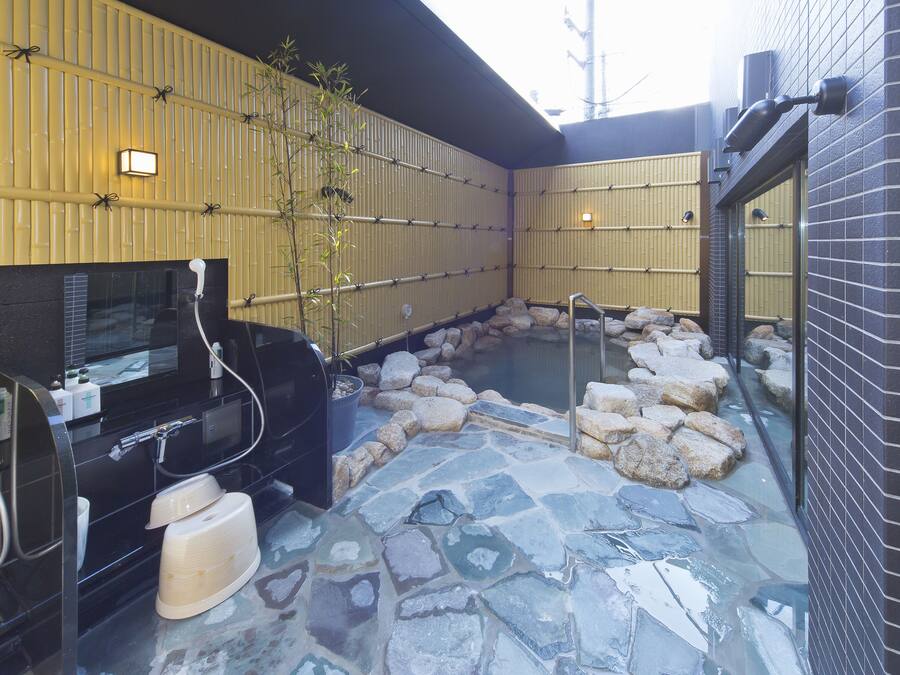 A traditional Japanese onsen with a stone hot spring surrounded by rocks, a black grill, and a bamboo wall.