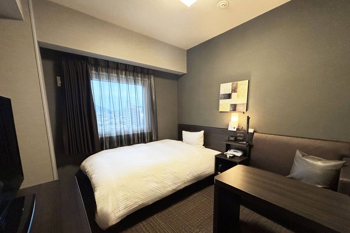A hotel room with a bed, a desk, a chair, a nightstand with a lamp, and a wall-mounted artwork.