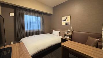 A hotel room with a bed, a desk, a chair, a nightstand, a lamp, and a window with curtains.