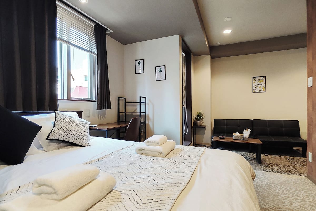 Hotellihuone, jossa on sänky, työpöytä, tuoli, sohva ja pieni pöytä, jonka päällä on kasvi.