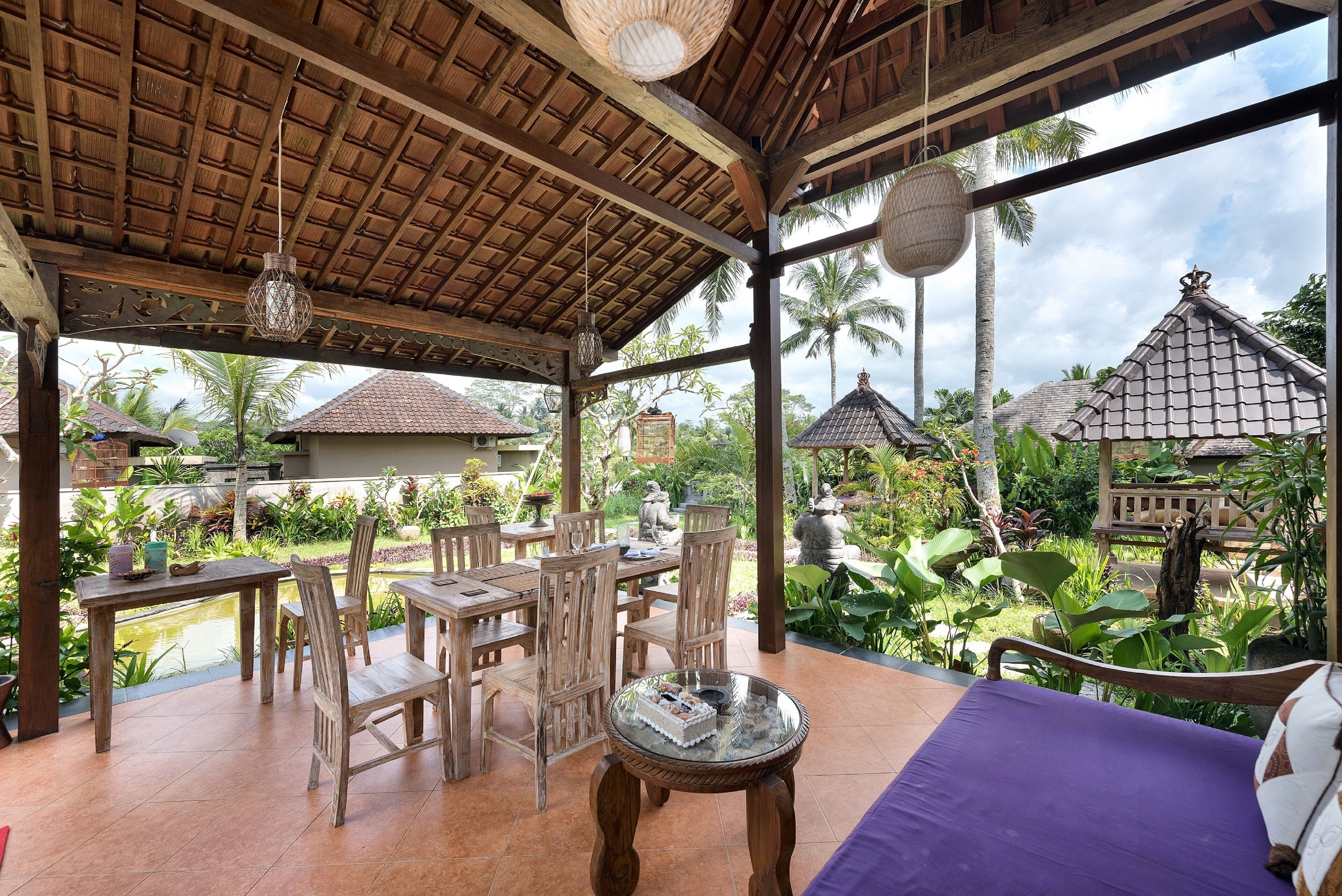 A covered outdoor seating area with wooden furniture, a glass table, and a view of a tropical garden with palm trees and a pond.