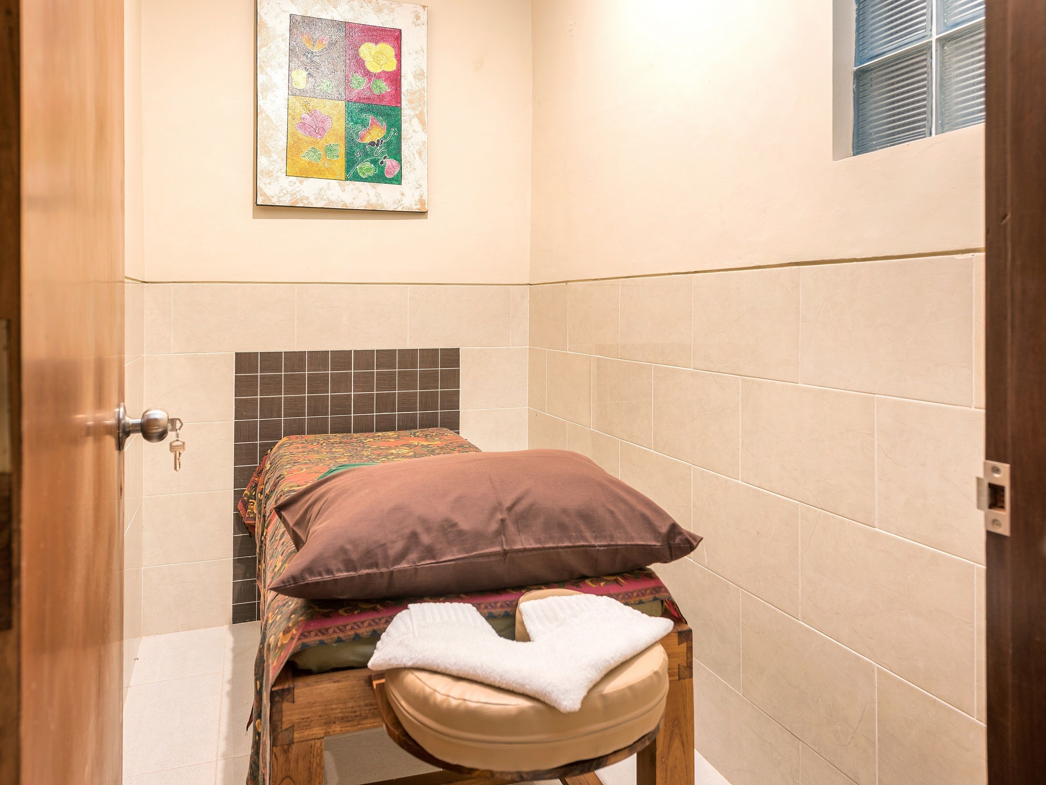 A small, cozy bathroom with a shower area, a wooden stool, a towel, and a colorful artwork on the wall.