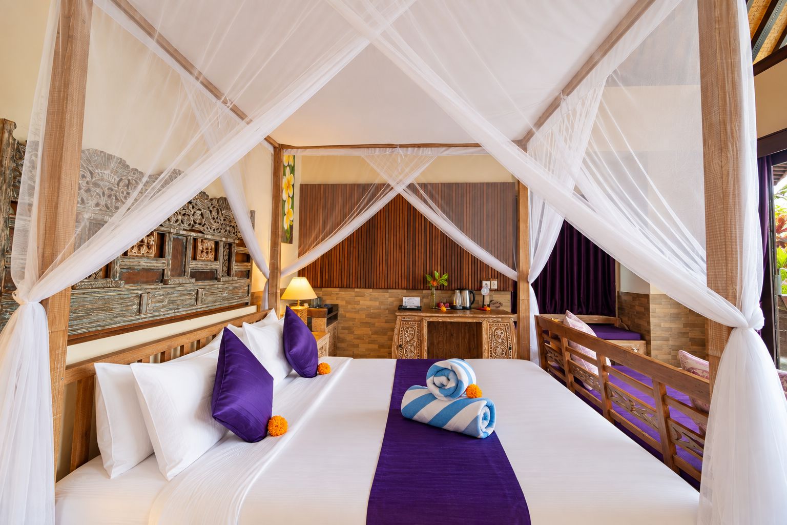 A room with a bed under a canopy, a wooden desk, a nightstand, and a view of the outside.