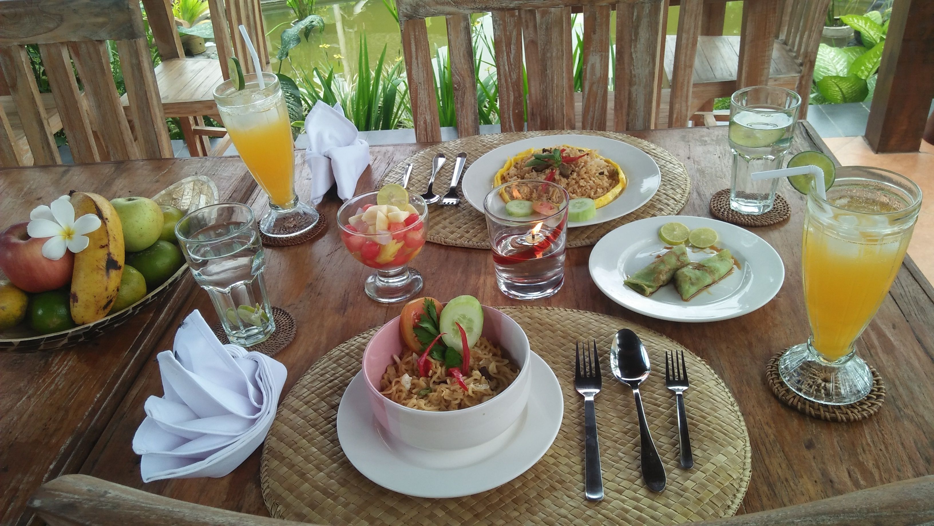 A wooden table set with various dishes, drinks, and fruits, suggesting a meal setup in an outdoor setting.