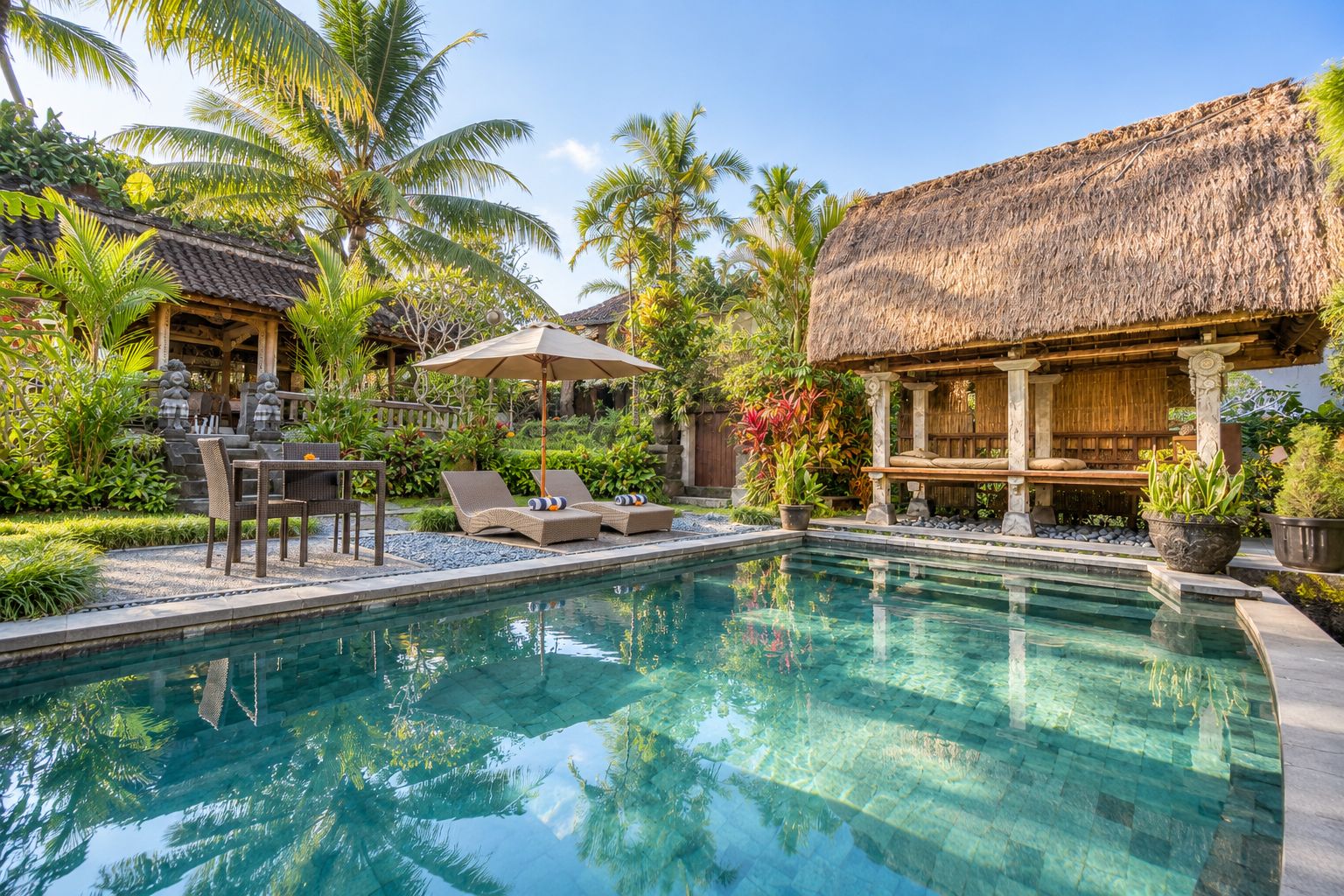 A clear swimming pool with lounge chairs and a table, surrounded by tropical plants and thatched-roof structures.
