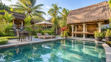 Piscina cristalina con sillas de descanso y una mesa, rodeada de plantas tropicales y estructuras de techo de paja.