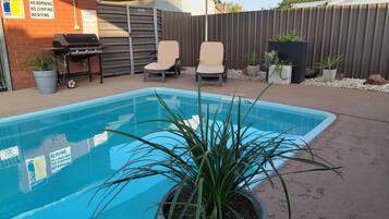 Un patio trasero con piscina, sillas de descanso, una parrilla y un área cercada con plantas en macetas.