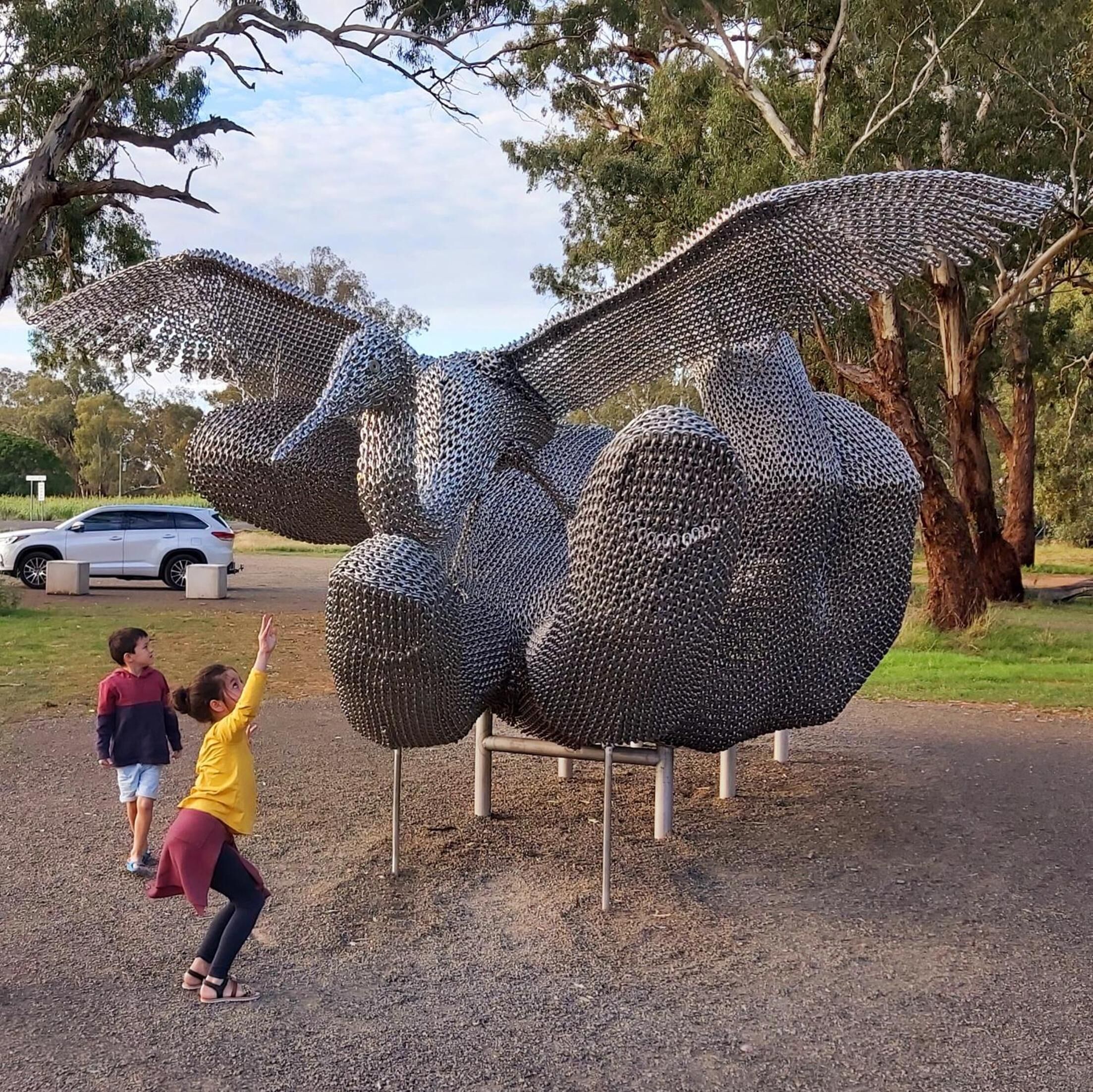 Una gran escultura de un animal hecha de cadenas, ubicada al aire libre con árboles y una carretera de fondo.