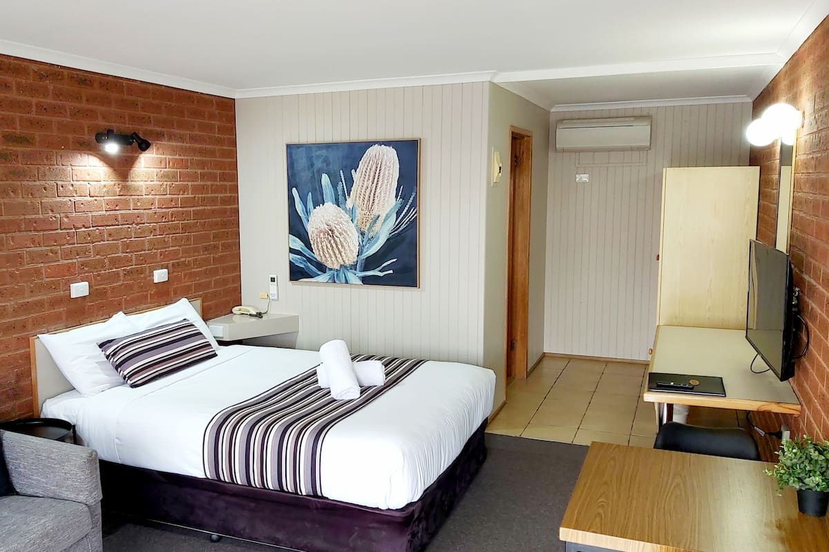A hotel room with a brick wall, a bed with striped bedding, a grey armchair, a wooden side table, a TV, and a painting of flowers.
