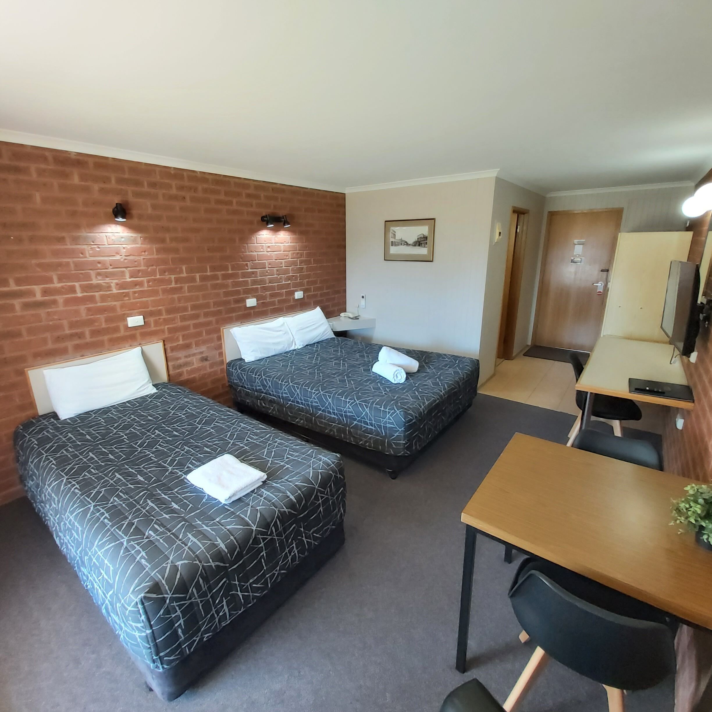 A hotel room with two beds, a desk, a chair, a TV, and a brick wall.