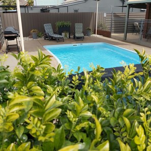 A rectangular swimming pool with a surrounding patio area, including two lounge chairs and potted plants.