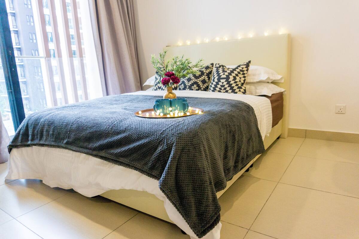 A neatly made bed with a dark blanket, a tray with flowers, and patterned pillows.
