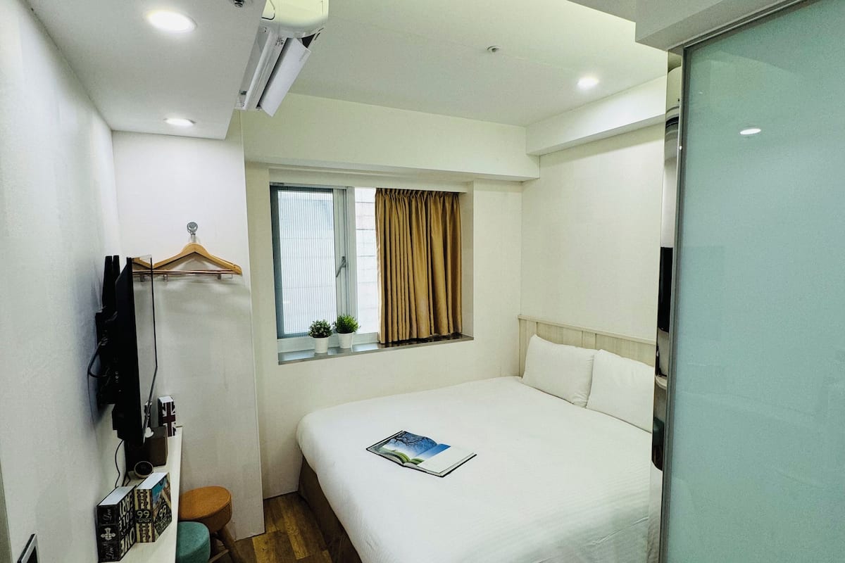 Une chambre d’hôtel comprenant un lit, une table de chevet, une fenêtre avec des rideaux, un petit tabouret et un téléviseur fixé au mur.