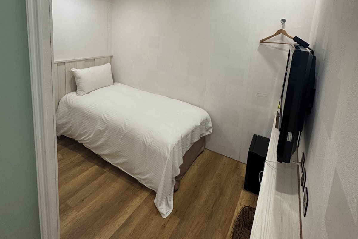 Une petite chambre à coucher, bien rangée, avec un lit simple, un plancher en bois, un climatiseur fixé au mur et une petite étagère sur laquelle est posé un objet.