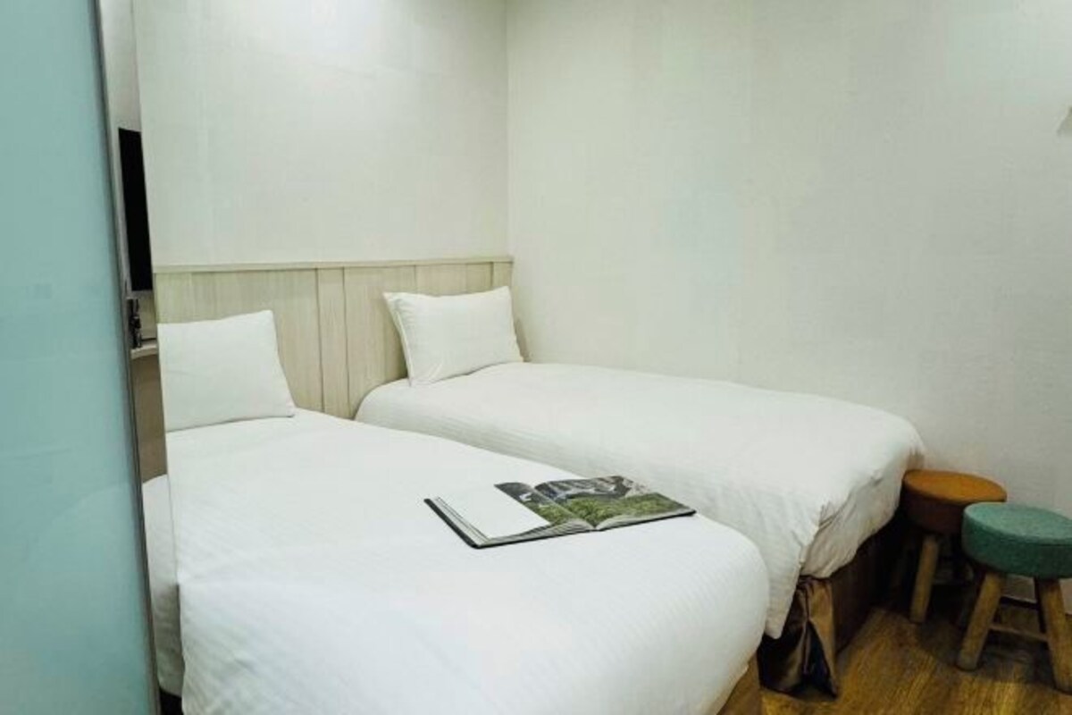 A small, simple room with two single beds, a small stool, and a magazine on one of the beds.