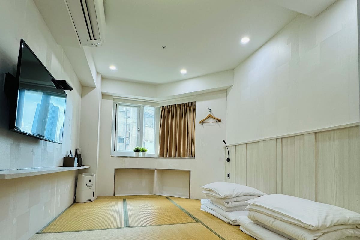 Une chambre d’hôtel avec un sol recouvert de tatamis, un lit avec du linge de maison blanc, un téléviseur fixé au mur, une fenêtre avec des rideaux et un radiateur fixé au mur.