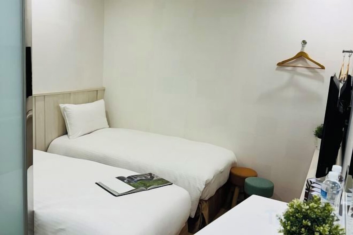 A small, compact room with two single beds, a small table with a magazine, and a hanging clothes rack.