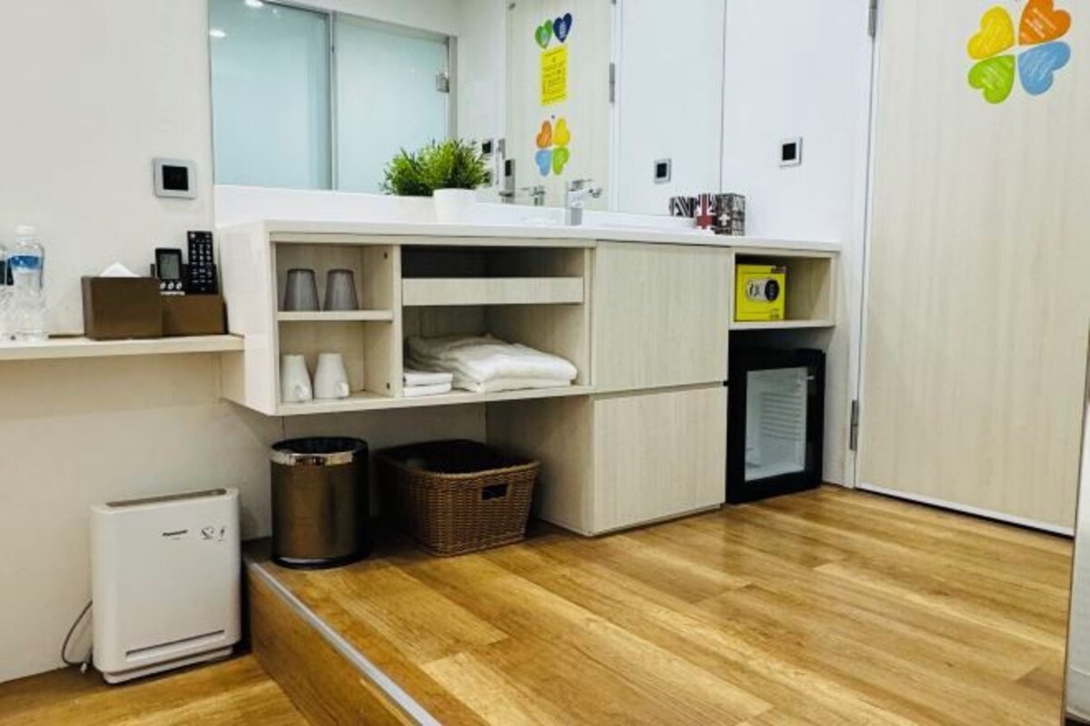 A modern bathroom with a wooden floor, a white sink, a mirror, and a small plant. There are shelves with toiletries and a trash bin.