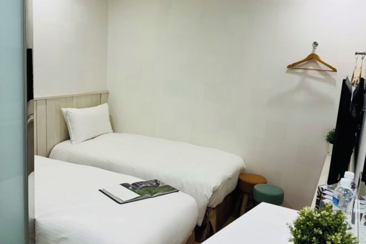 A small, neatly arranged bedroom with two beds, a small stool, a table with toiletries, and a hanging clothes rack.