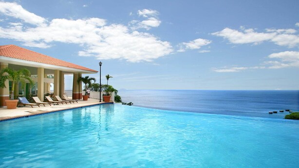 A pool with lounge chairs and potted plants, overlooking a clear blue sea.