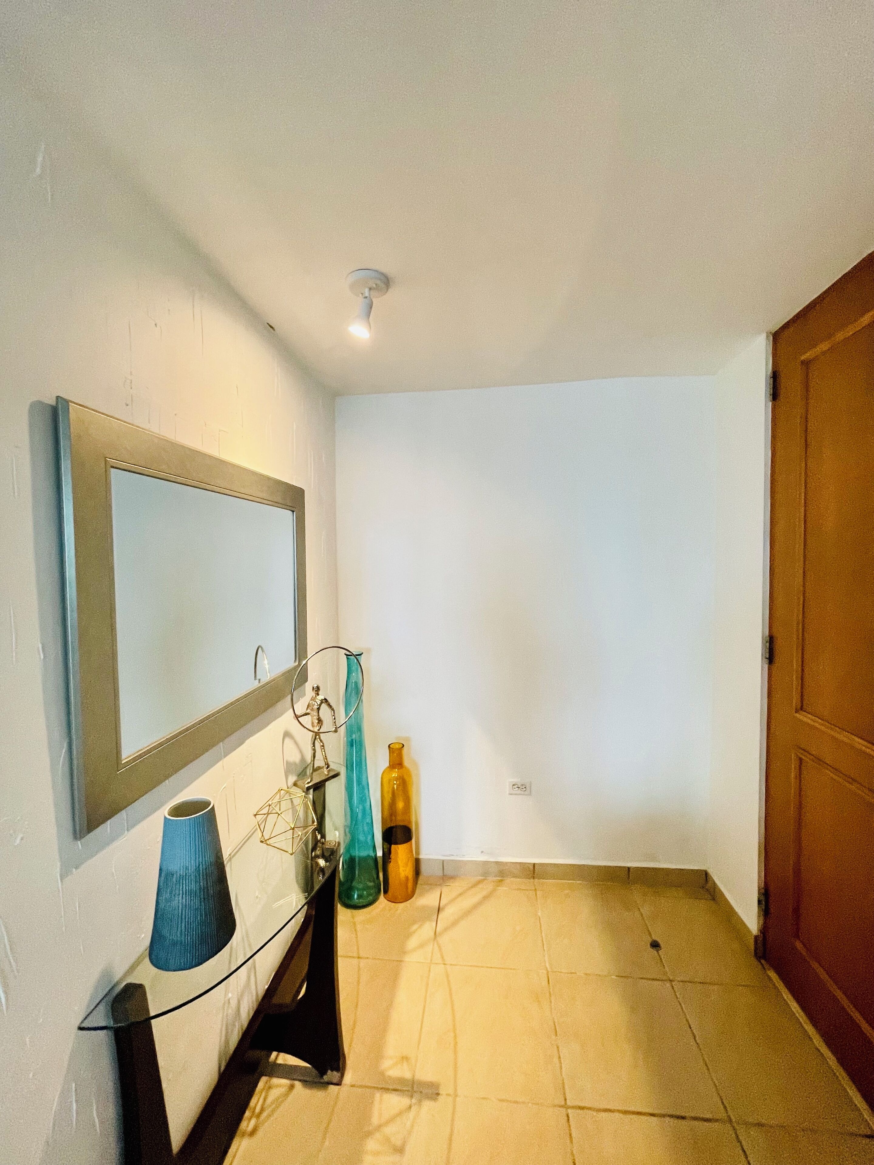 Una habitación pequeña y luminosa, con un espejo, un florero y una botella en el suelo.