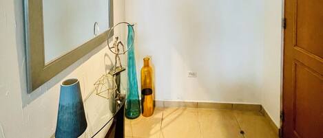 Una habitación pequeña y luminosa, con un espejo, un florero y una botella en el suelo.