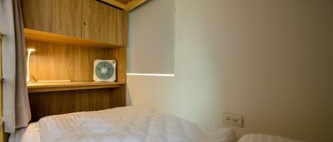 Una habitación pequeña y acogedora con cama, un estante de madera con un ventilador y una ventana.
