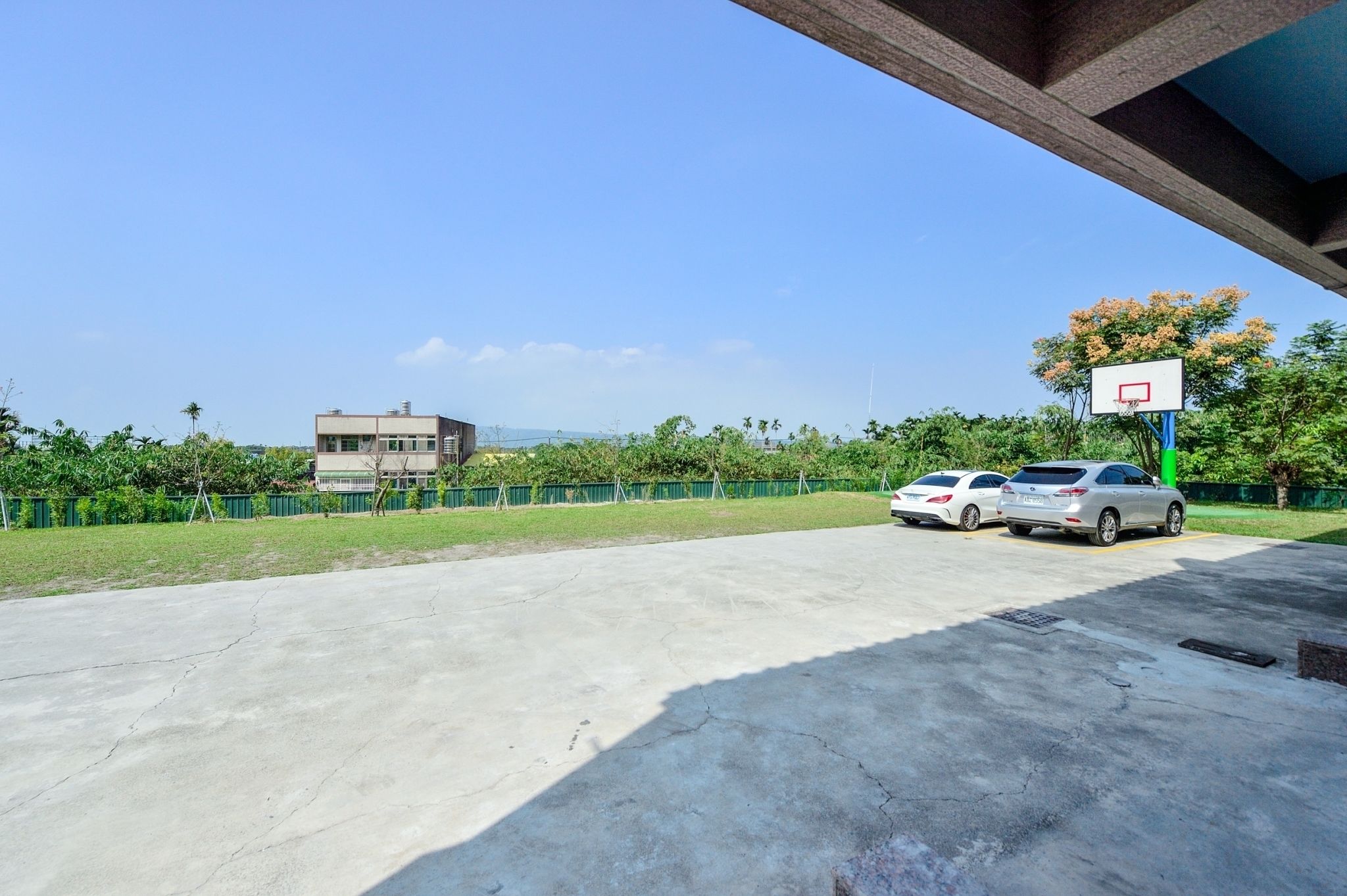 A parking area with two cars, a basketball hoop, and a building in the background.