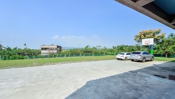 A parking area with two cars, a basketball hoop, and a building in the background.
