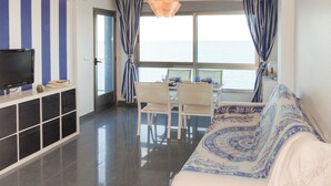 Uma sala de estar moderna com sofá, mesa de jantar e cadeiras. Uma televisão está montada na parede, e há vista para o mar através de grandes janelas.