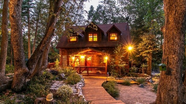 Une cabane dotée d’une terrasse en bois donnant sur un porche, le tout entouré d’arbres et bordé d’un sentier en pierre.