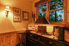 Een gezellige kamer met een houten dressoir, een lamp, ingelijste foto’s en een raam met uitzicht op bomen.