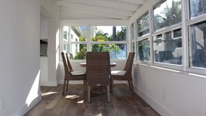 A sunlit room with a table and chairs, large windows, and a view of greenery outside.