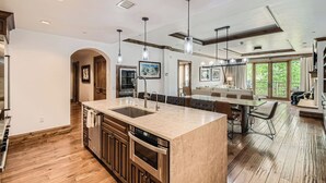 A modern kitchen with a large island, wooden cabinets, and a built-in oven. There is a dining area with a long table and chairs, and a seating area with a sofa and a coffee table.
