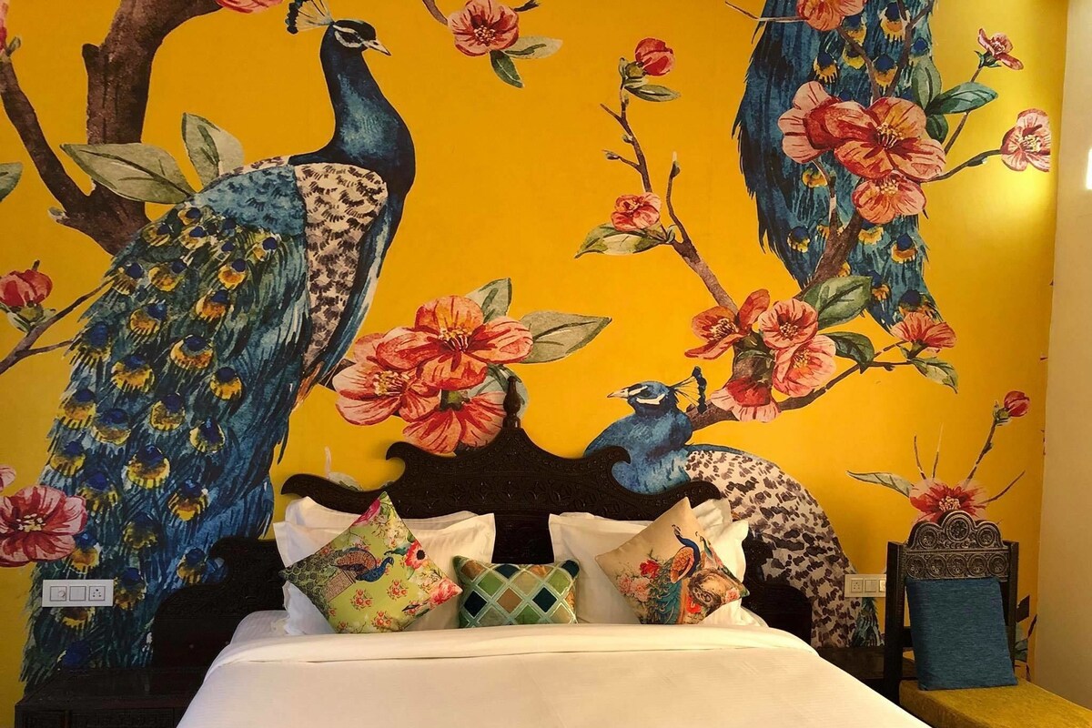 A bed with a headboard, multiple pillows, and a wall with a peacock and floral mural.