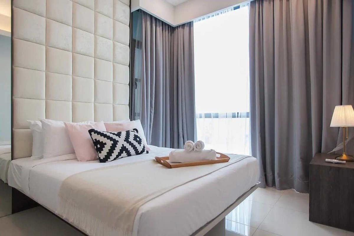A modern hotel room with a large bed, a tufted headboard, grey curtains, and a bedside table with a lamp.