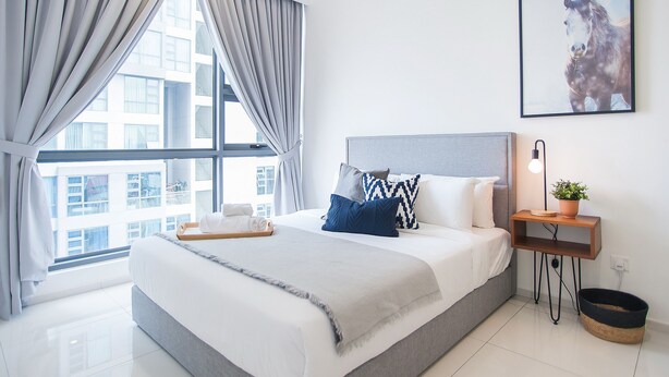 Ein modernes Hotelzimmer mit einem großen Bett, einem Nachttischchen mit Lampe, einem gerahmten Bild an der Wand und Blick auf die Stadt durch das Fenster.