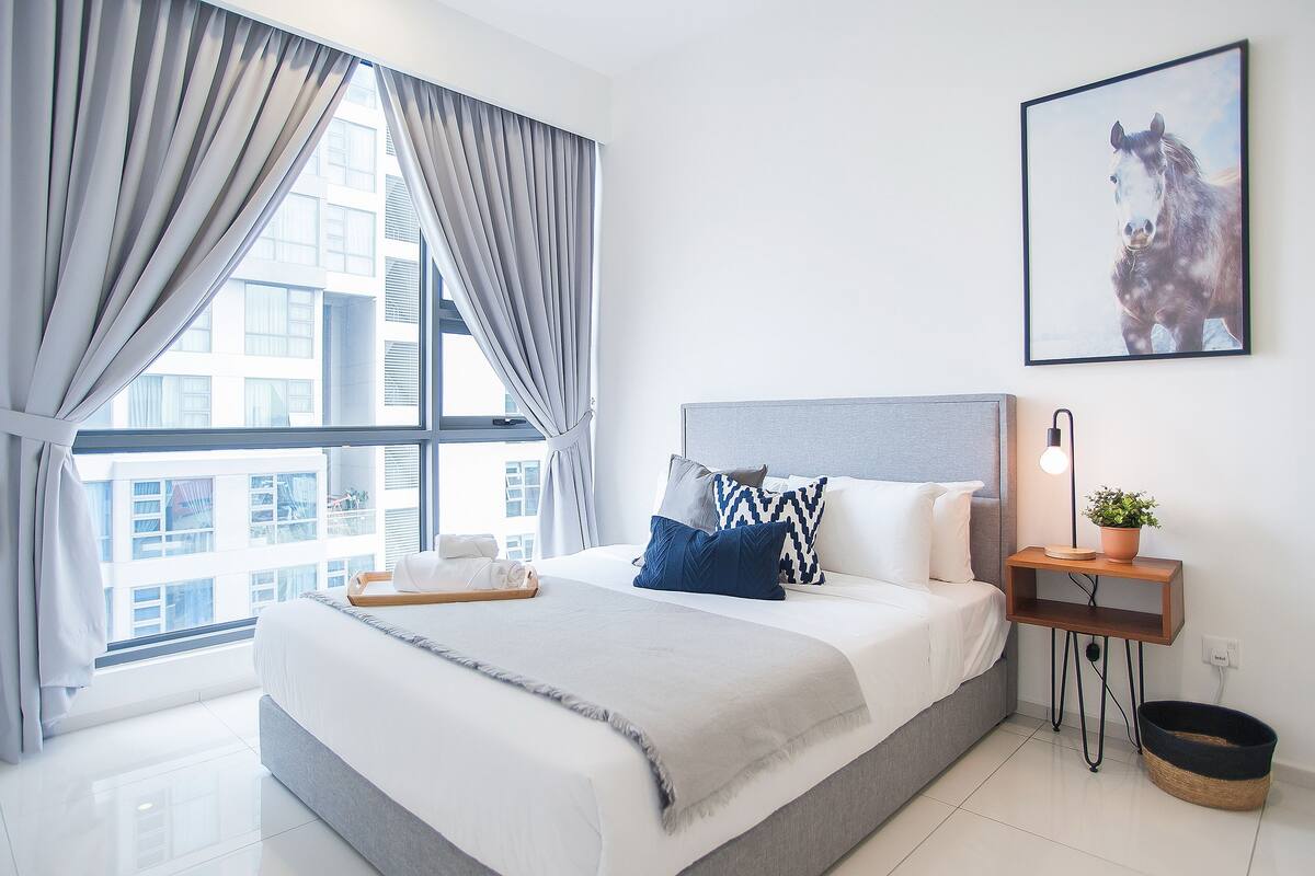 A modern hotel room with a large bed, a bedside table with a lamp, a framed picture on the wall, and a view of the city through the window.