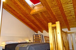 A room with a sloped wooden ceiling, a bed with a patterned blanket, a small table with a lamp, and a window with a red frame.