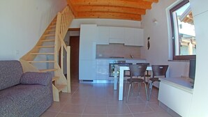 A living room with a sofa, a dining area with chairs and a table, a kitchen with cabinets, and a wooden staircase. - La Quiete 58 Holideal (Tremosine)