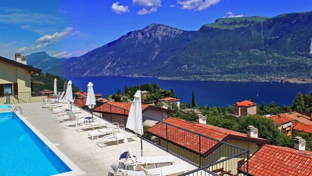 A swimming pool with lounge chairs and umbrellas, surrounded by buildings with red-tiled roofs, set against a backdrop of mountains and a lake. - La Quiete 58 Holideal (Tremosine)