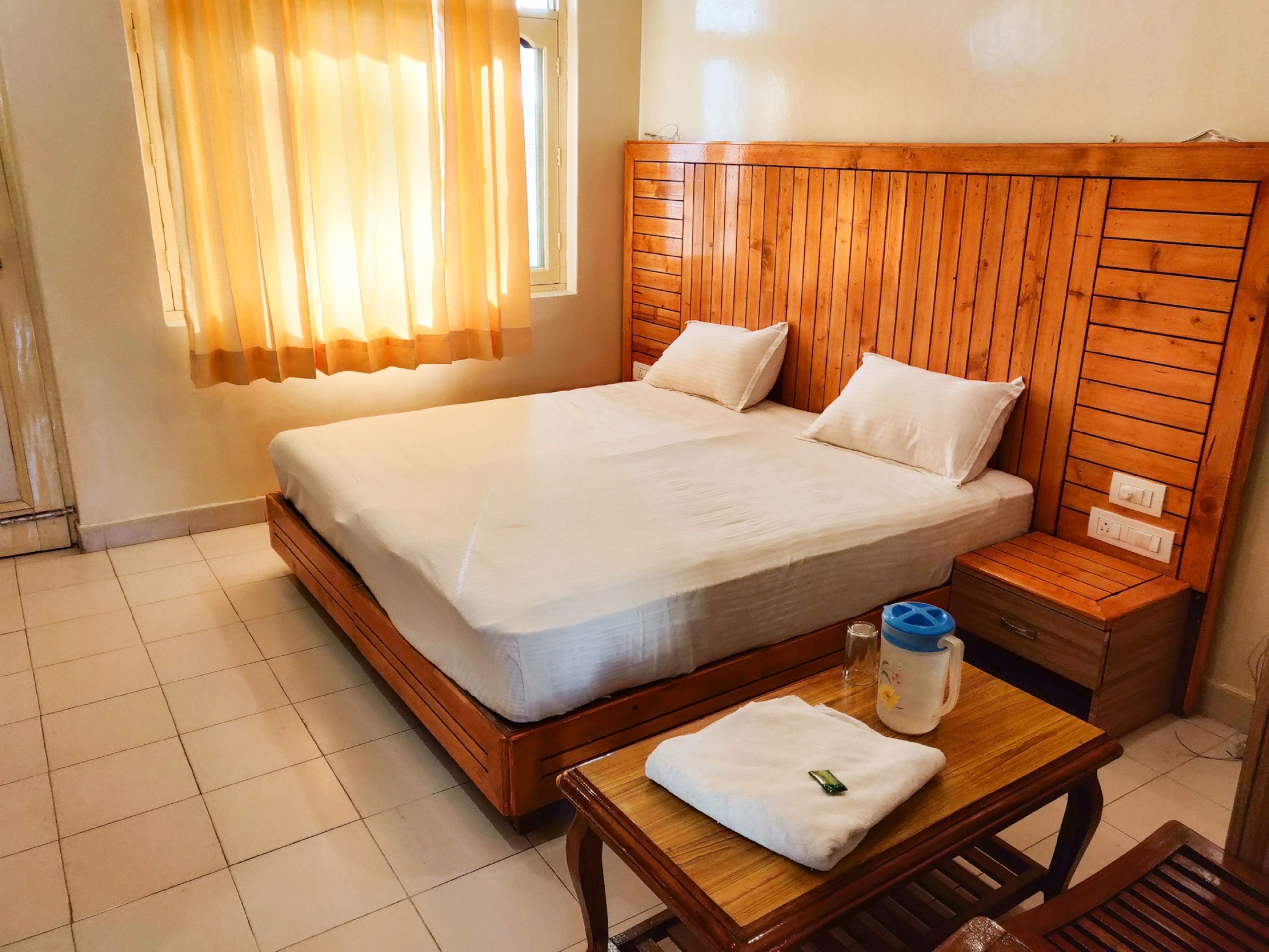 A hotel room with a wooden bed, a wooden nightstand, a bedside table with a towel and a water bottle, and a window with curtains.