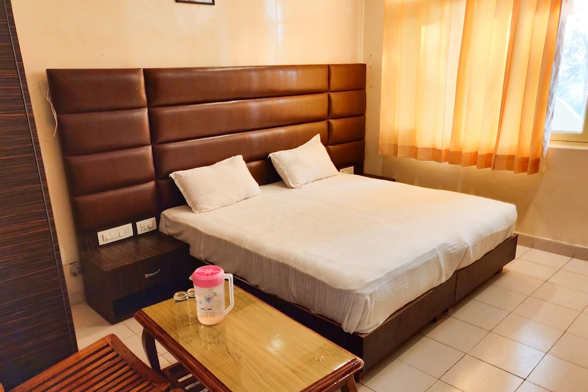 Una habitación de hotel con una cama grande, una mesita de noche de madera, una pequeña mesa con una taza rosa y una ventana con cortinas.