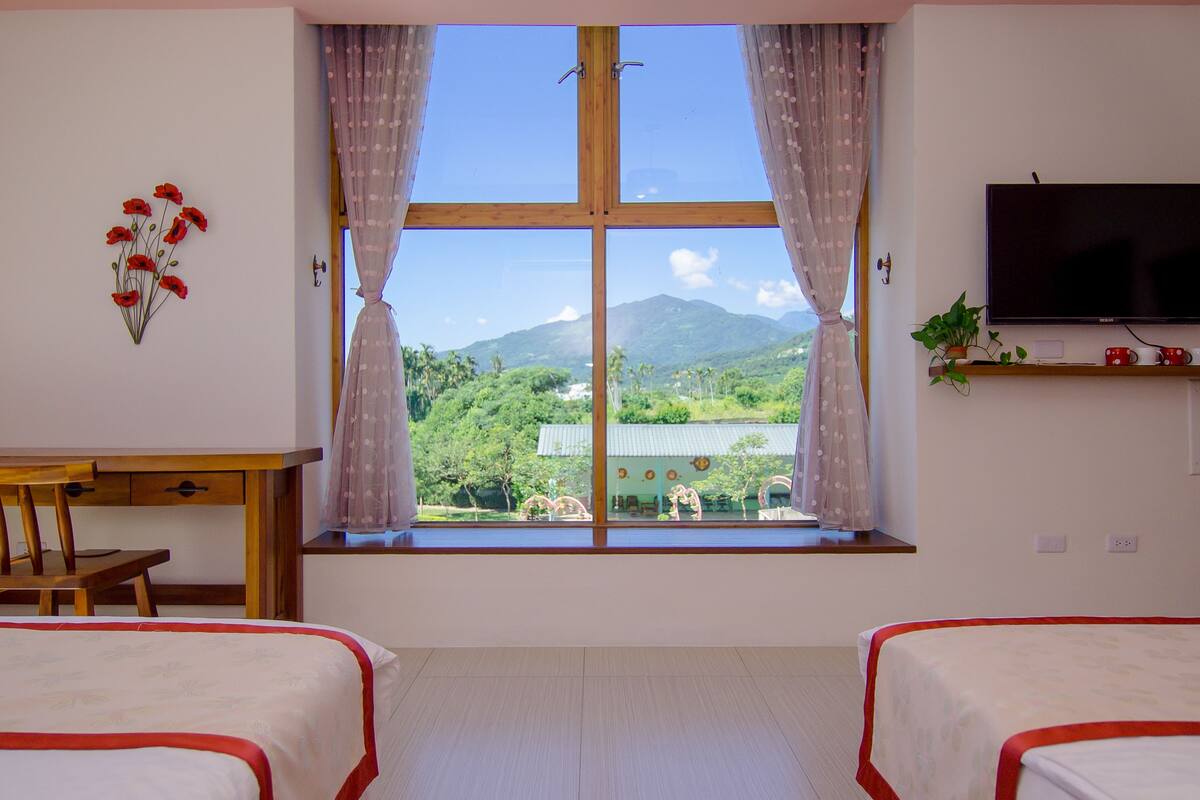 A room with two beds, a desk, a TV, and a window with a view of mountains and greenery.