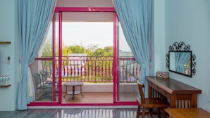A room with a sliding glass door leading to a balcony with a view of greenery and a fence. There is a small table and chairs on the balcony, and a wooden desk with drawers in the room. - XinFu Traveler Inn (Beinan)