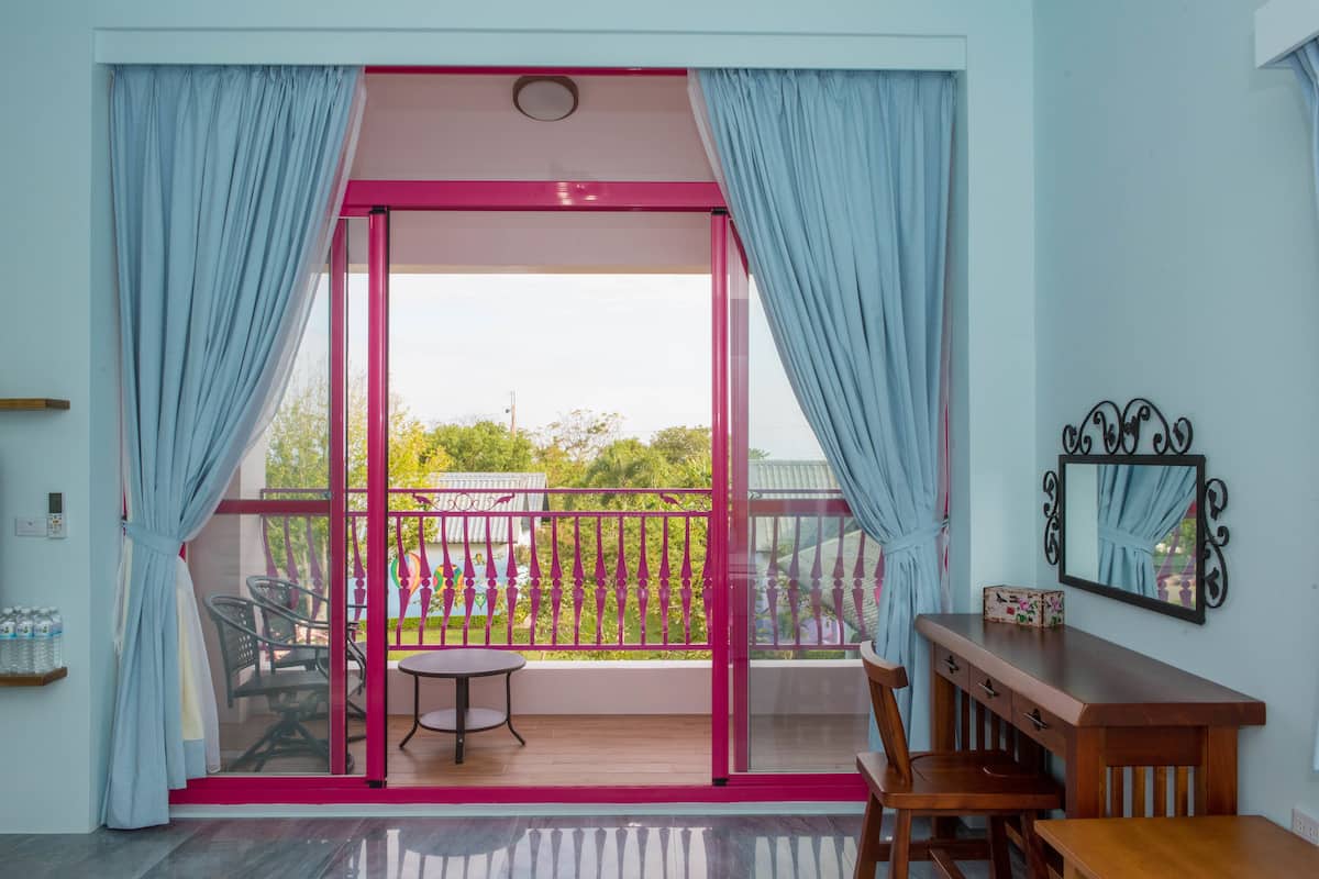 A room with a sliding glass door leading to a balcony with a view of greenery and a fence. There is a small table and chairs on the balcony, and a wooden desk with drawers in the room.