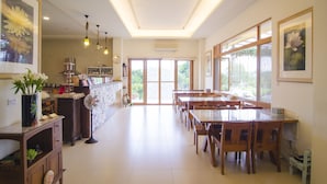 A spacious dining area with multiple wooden tables and chairs, a kitchen area with a refrigerator and stove, and large windows providing natural light and views of the outdoors. - XinFu Traveler Inn (Beinan)