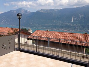 A balcony with a tiled floor, a metal railing, and a street view with buildings and mountains in the background.
