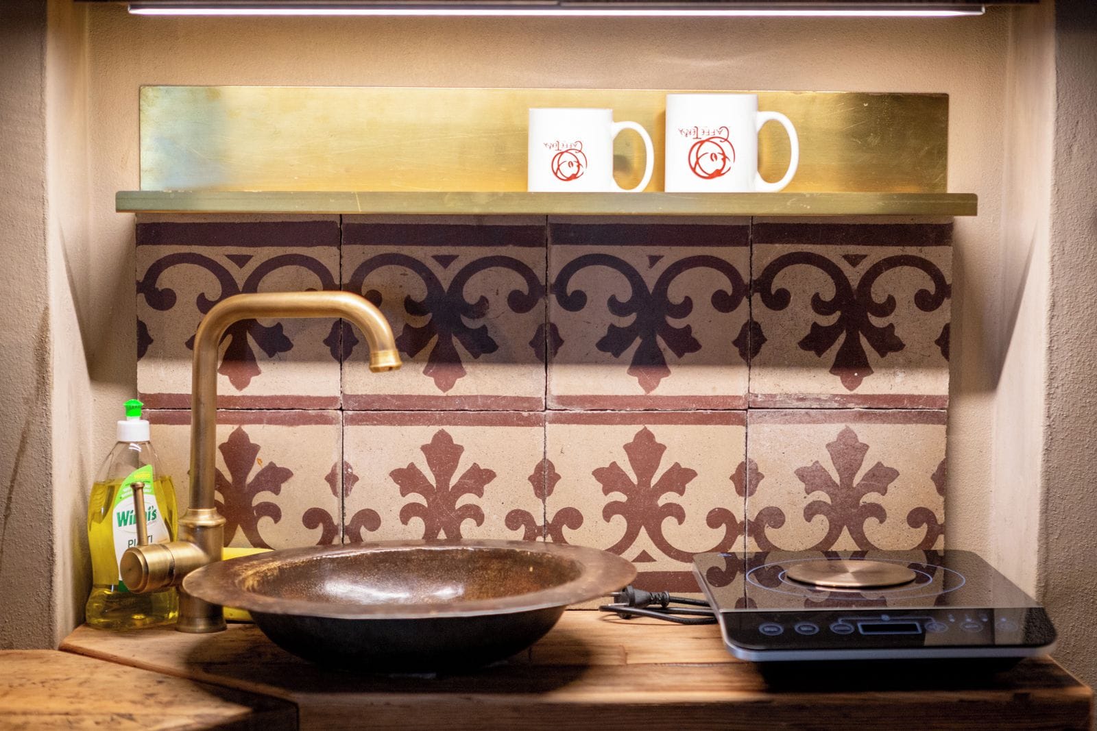 Une cuisine avec un évier en céramique marron, un plan de travail en bois, une bouteille de liquide vaisselle, une cuisinière à gaz et un crédence ornée d’un motif décoratif en carreaux.