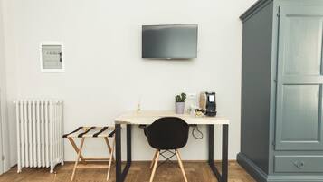 A room with a wooden floor, a white radiator, a small table with a chair, a television mounted on the wall, and a cabinet.